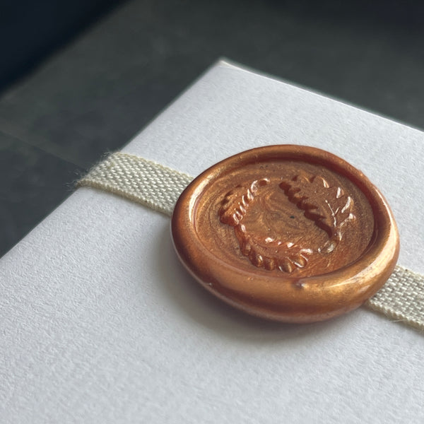 A wax seal stamp with an oak leaf design, using a rose wood handle, pressed onto a white card with a linen ribbon.