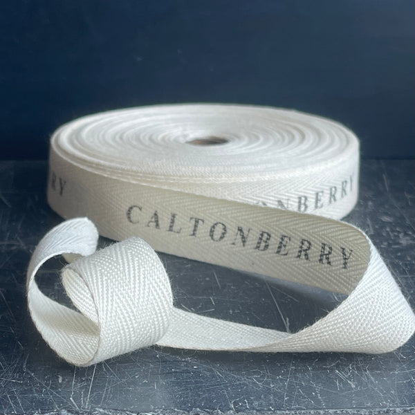 A roll of natural cotton ribbon with the text 'CALTONBERRY' printed on it, displayed on a dark surface.