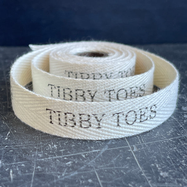 natural cotton herringbone ribbon with the words 'TIBBY TOES' printed on it, placed on a textured surface.