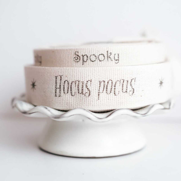 Rolls of beige cotton ribbon with the words 'Spooky' and 'Hocus pocus' printed on them, displayed on a white stand.