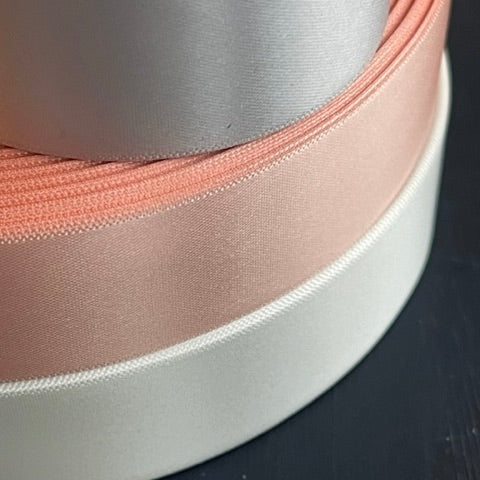 Rolls of satin ribbon in three different shades: silver, white, and pink, with a black print. The ribbon is 25mm wide and appears to be silky in texture.